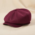Casquette gavroche bordeaux posée sur tissu crème, photo studio produit fond chaud